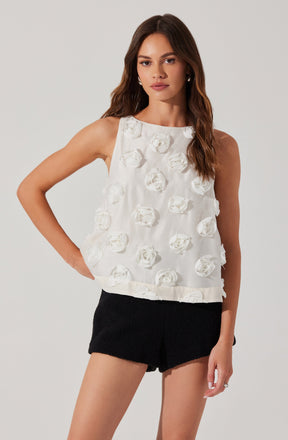A young woman is wearing a sleeveless, white top decorated with 3D floral embellishments. The top has a round neckline and a loose fit. She pairs it with black shorts that have a textured fabric.