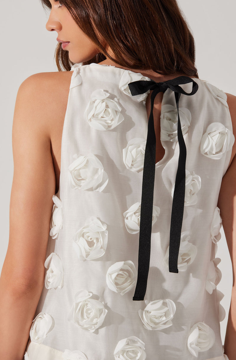 A woman is wearing a sleeveless white top decorated with three-dimensional white rose appliqués covering the entire fabric. The top features a black bow tied at the back of the neckline with long black ribbons hanging down.