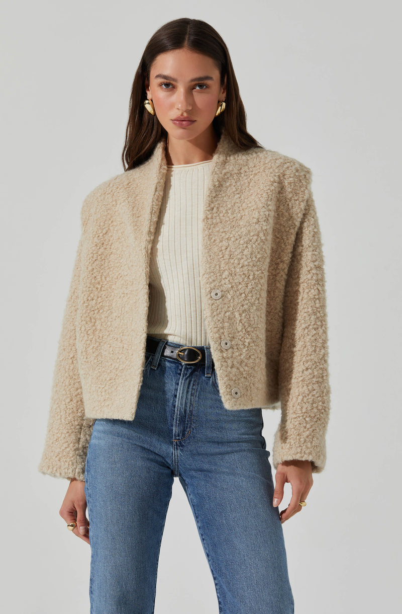 A woman is wearing a cream-colored, textured, fleece or teddy-style jacket with a collarless design and large buttons down the front. She has paired it with a white, ribbed, crew-neck top and high-waisted blue jeans.