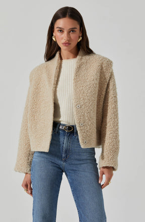 A woman is wearing a cream-colored, textured, fleece or teddy-style jacket with a collarless design and large buttons down the front. She has paired it with a white, ribbed, crew-neck top and high-waisted blue jeans.