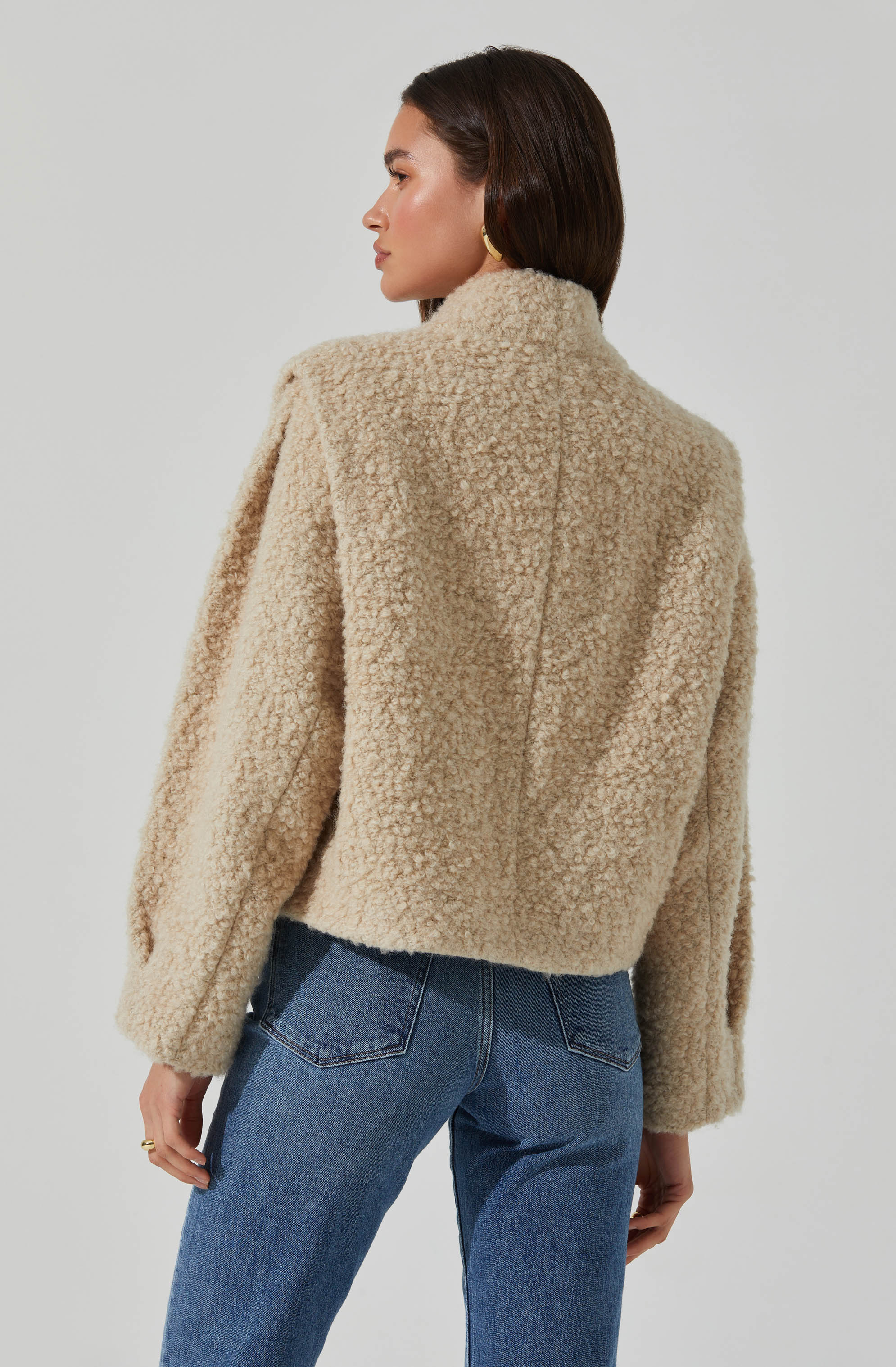A woman is wearing a beige, textured, and plush sherpa fleece jacket with a high collar, paired with medium-wash blue jeans.