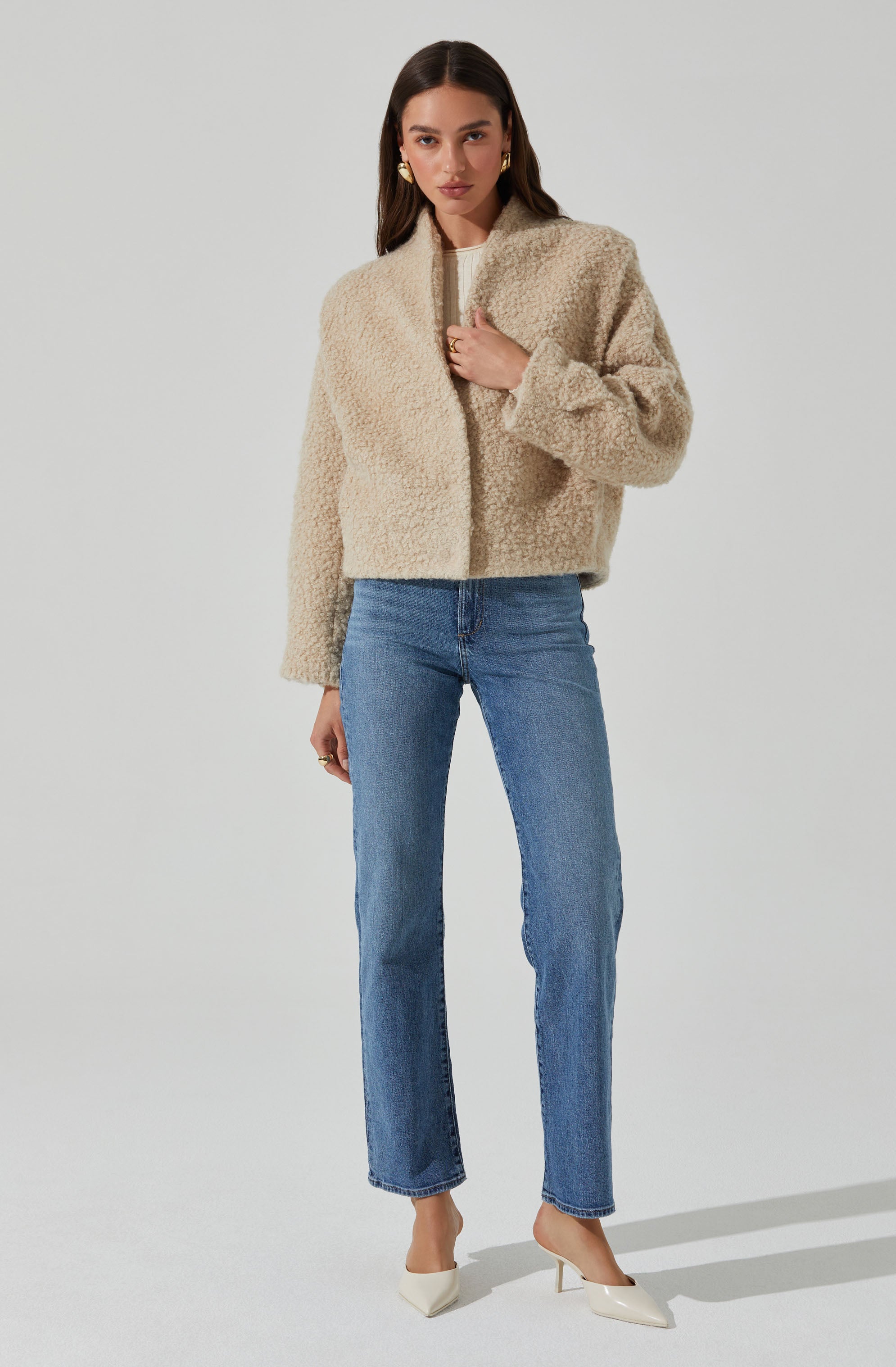 A woman is wearing a beige, textured, zip-up fleece jacket with a slightly cropped, boxy fit. She pairs it with high-waisted blue jeans that have a straight-leg cut. She completes her outfit with white pointed-toe heels. She also has gold earrings and rings as accessories.