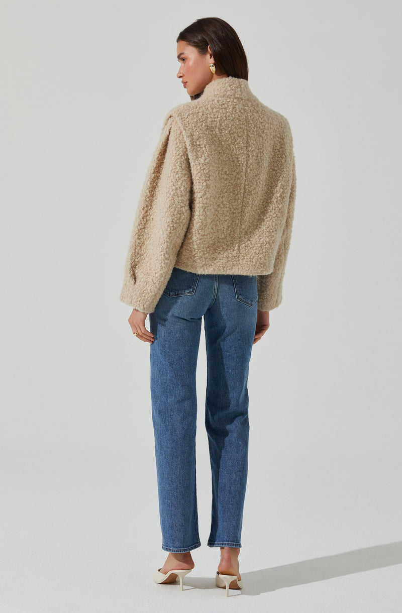 A woman is wearing a beige, textured, fleece jacket with a collar. She pairs it with high-waisted blue jeans and white heeled shoes. She has dark, shoulder-length hair and gold earrings.