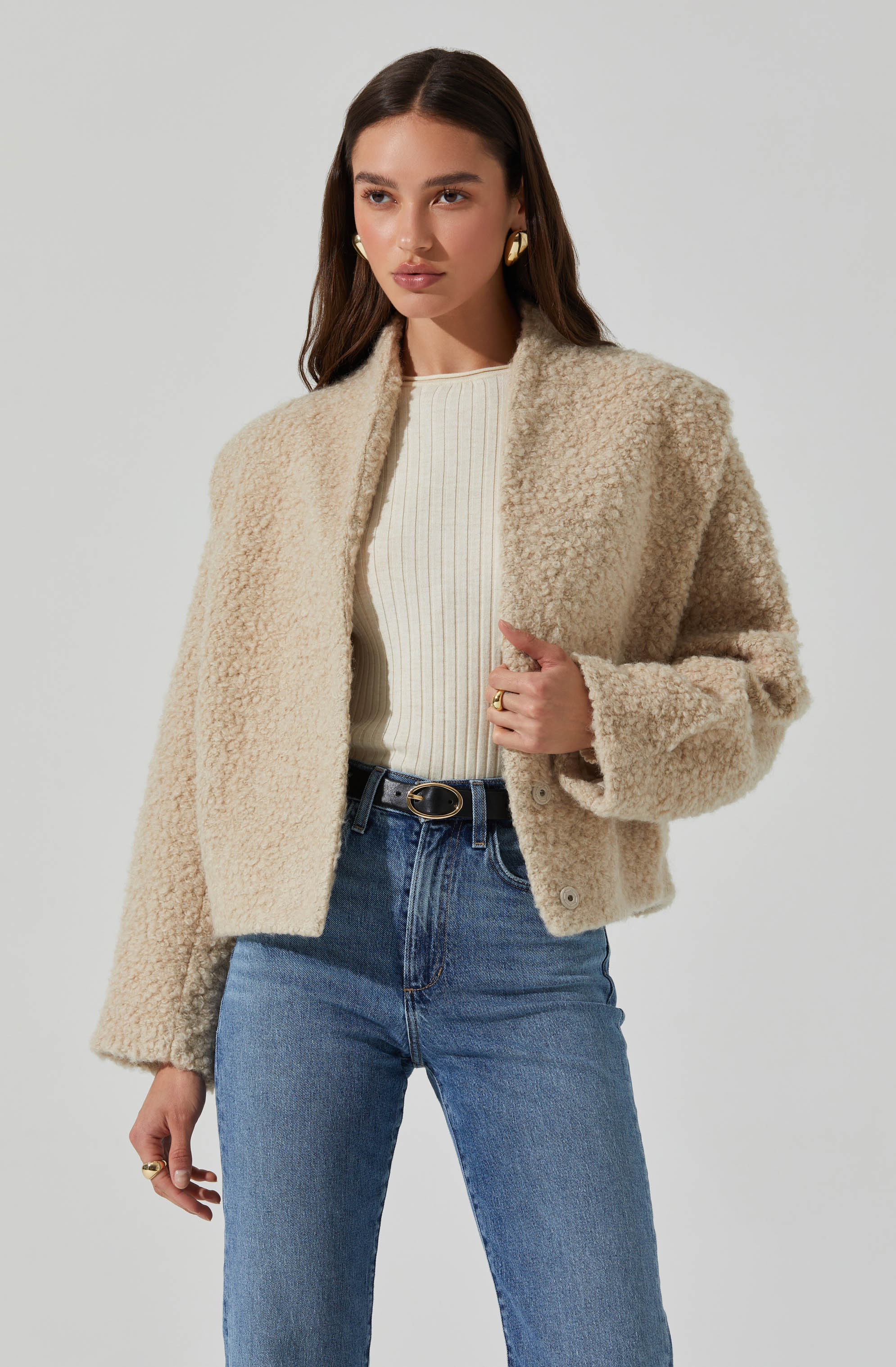 A woman is wearing a beige, textured, fleece or teddy-style jacket with a round neckline, paired with a cream-colored ribbed knit top and high-waisted blue jeans. The outfit is accessorized with gold earrings and rings, and she has dark, wavy hair.
