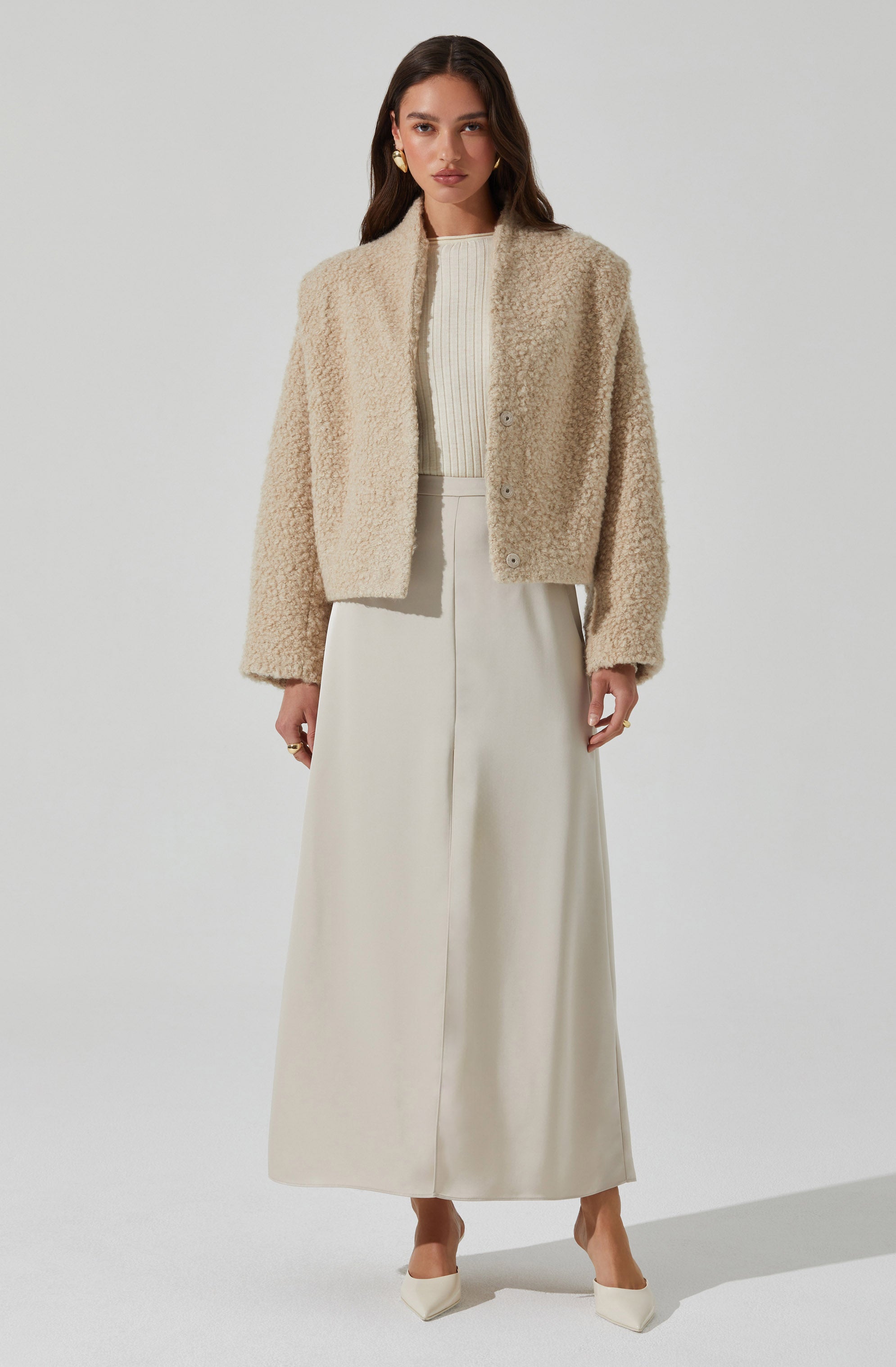 A woman is wearing a cream-colored, plush, teddy bear coat with a high collar and buttons down the front. Underneath, she has on a ribbed, cream-colored top. She pairs these with a long, flowing, cream-colored skirt and pointed-toe heels that match her outfit. She also has gold earrings and rings as accessories.