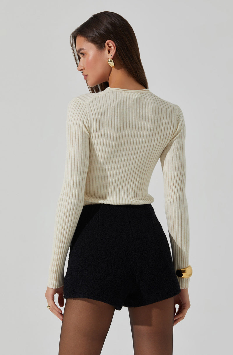 A woman is wearing a fitted, long-sleeve, ribbed sweater in a light cream or beige color. She pairs it with high-waisted black shorts and accessorizes with gold earrings, a gold bracelet, and rings. Her dark brown hair is styled straight, and she has a neutral expression.