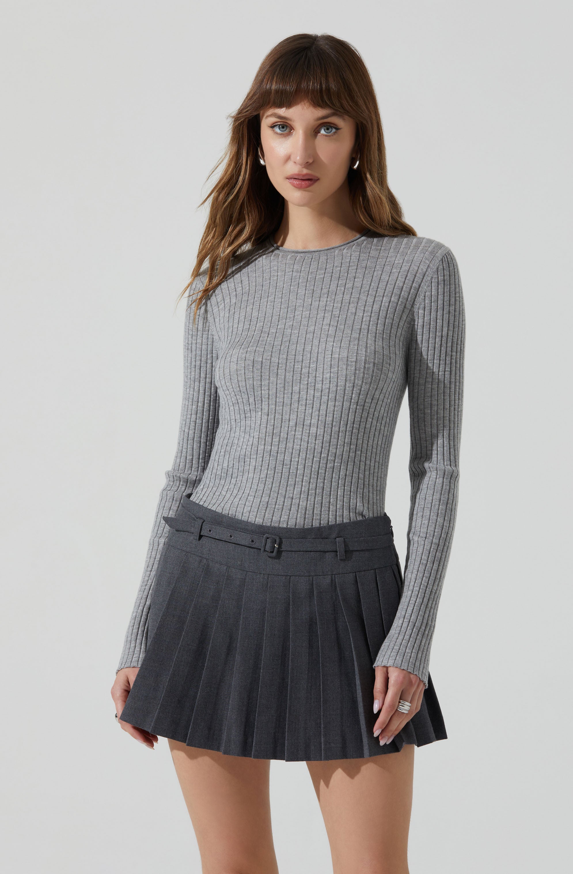A woman is wearing a fitted, long-sleeved, gray ribbed sweater with a round neckline. She pairs it with a high-waisted, dark gray pleated skirt that has a matching belt.