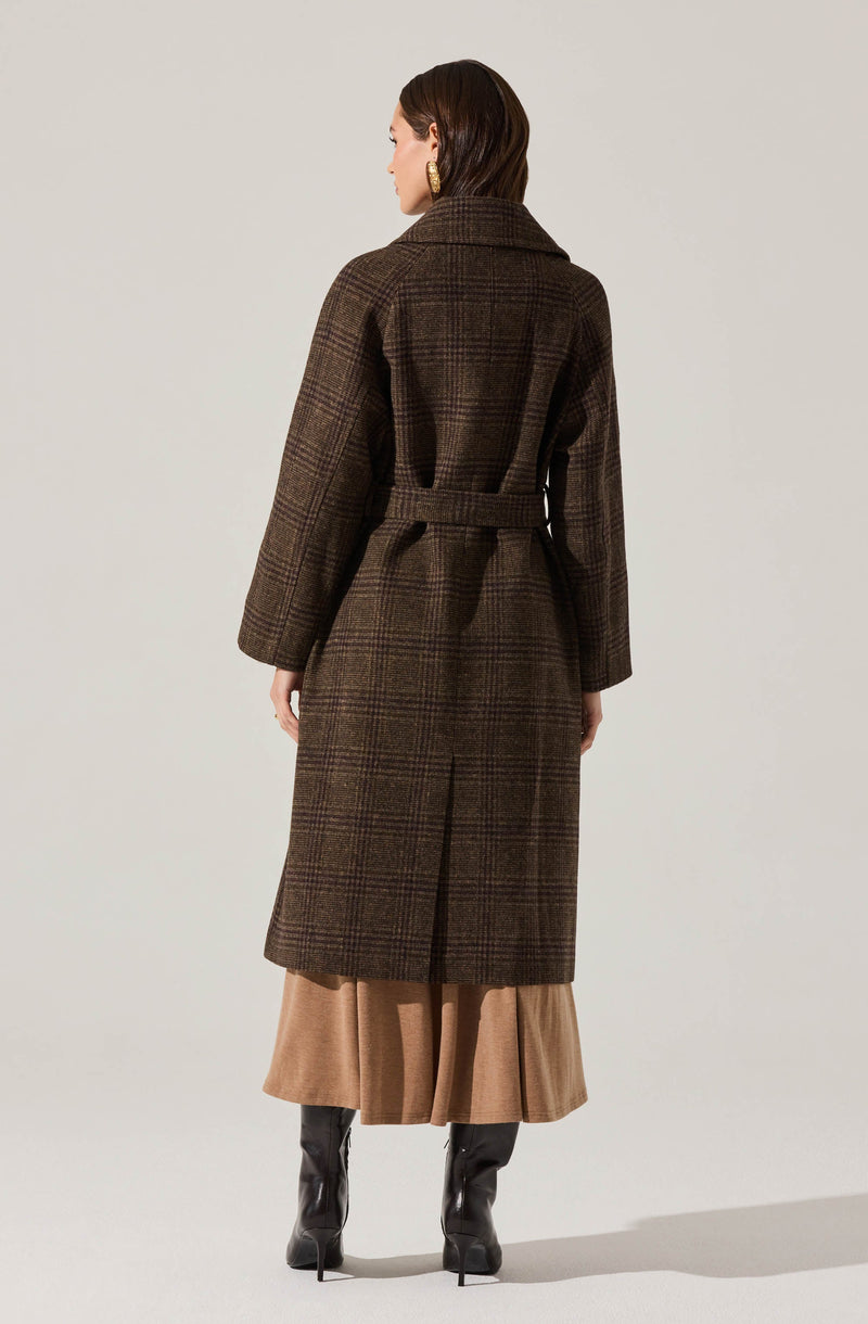 A woman is wearing a long, dark brown plaid coat with a belt at the waist and a wide collar. The coat has a classic checkered pattern and falls below the knees. Underneath, she is wearing a beige or tan pleated skirt that reaches mid-calf. She is also wearing tall black heeled boots. Her hair is dark brown, styled straight, and she is wearing gold earrings.