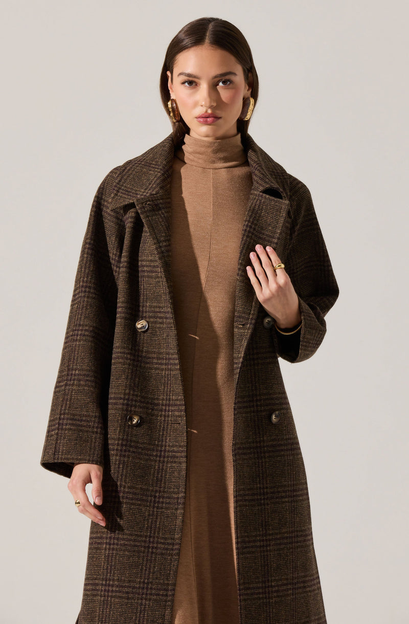A woman is wearing a long, double-breasted coat with a dark brown and green plaid pattern. Underneath, she has on a high-neck, camel-colored dress or top. She is accessorized with gold hoop earrings and a ring on her left hand.