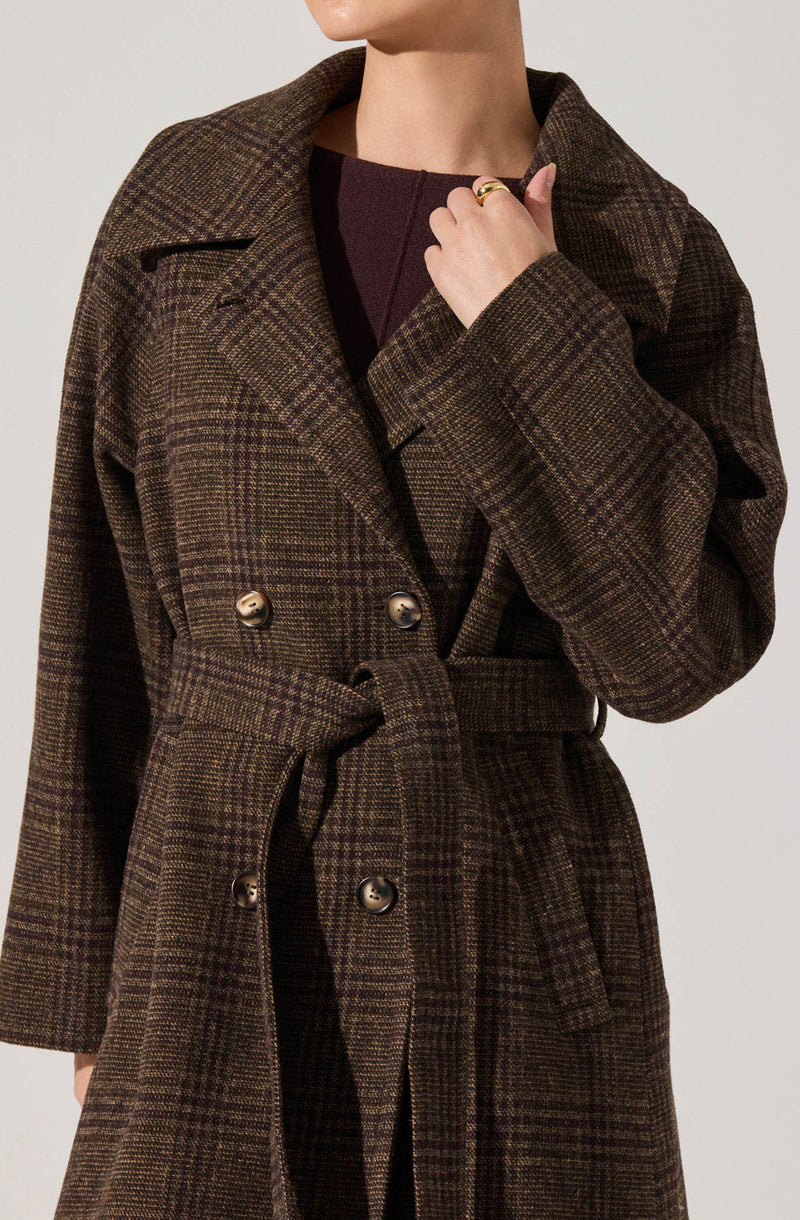 A person is wearing a long, brown plaid coat with a waist tie and large buttons. Underneath, they have on a dark top, possibly a sweater. The coat features a classic checkered pattern in shades of brown and green.