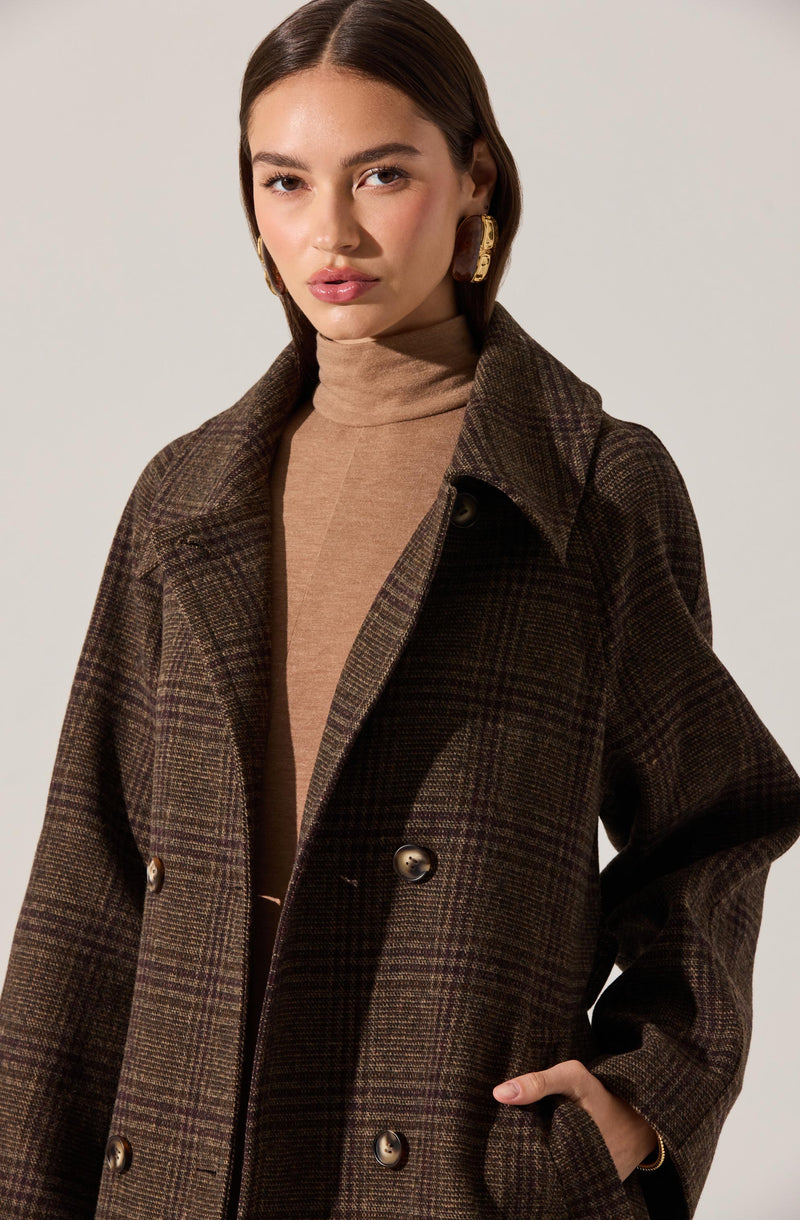 A woman is wearing a light brown turtleneck sweater underneath a dark plaid double-breasted coat with wide lapels. She has large gold earrings and her hair is styled sleekly.