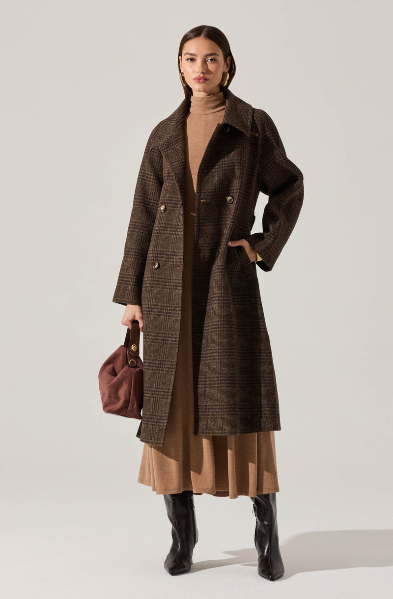 A woman is wearing a long, dark brown plaid coat with a double-breasted design and wide lapels. Underneath, she has on a beige turtleneck sweater. She is also wearing a beige pleated skirt that reaches mid-calf and pairs with black knee-high boots. She holds a small brown handbag with a gold clasp and hardware, and her accessories include large, gold hoop earrings.
