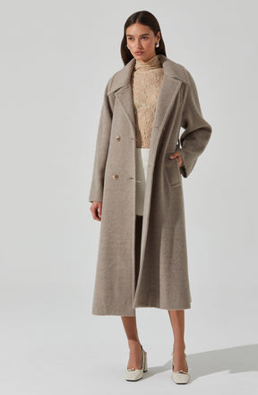 The woman is wearing a long, light beige or taupe-colored wool coat with a wide collar and two buttons, styled open. Underneath, she has on a cream-colored, high-neck, textured sweater. She is also wearing white or cream-colored wide-leg pants and white high-heeled shoes.