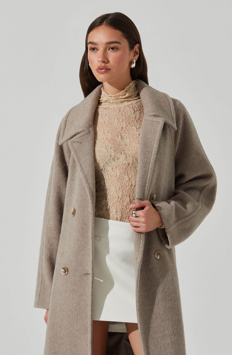 A woman is wearing a long, beige wool coat with large lapels and double-breasted buttons. Underneath, she has on a light, textured high-neck top in a neutral tone, and a white, high-waisted skirt that is above knee length. She has dark, straight hair and is accessorizing with earrings and a ring.