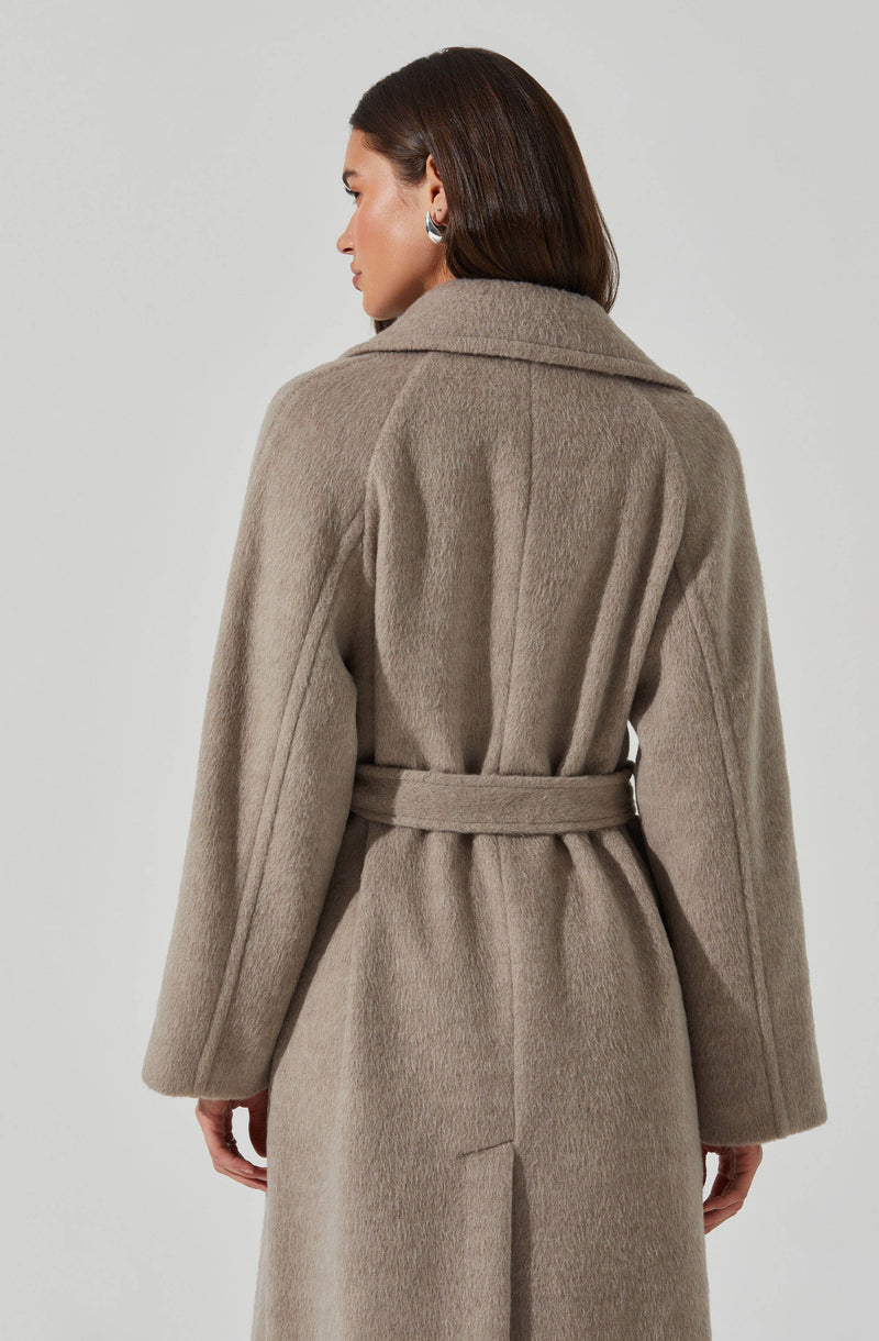 A woman is wearing a long, beige, wool coat with a belted waist. The coat has a wide collar, full-length sleeves, and a slit at the back.