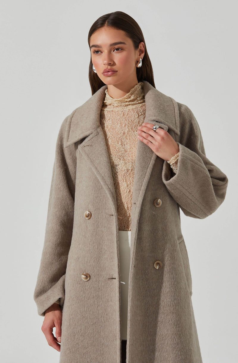 A woman is wearing a light beige, double-breasted, long wool coat with large lapels. Underneath, she has on a textured, high-neck top in soft pastel colors. She is accessorizing with large, round, silver earrings and a silver ring on her right hand.