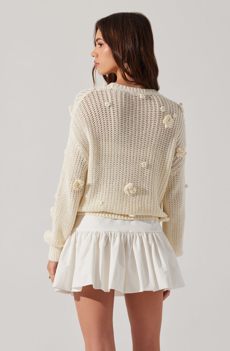 A woman is wearing a cream-colored, oversized knit sweater with textured pom-pom details scattered throughout. She pairs it with a high-waisted, white, ruffled mini skirt.