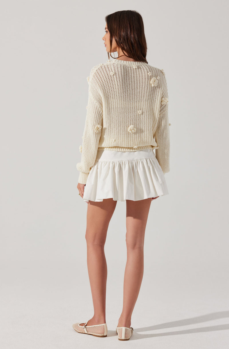 A woman is wearing a cream-colored, chunky knit sweater with textured pom-pom details scattered across the fabric. She pairs it with a white, ruffled short skirt and beige, open-toed shoes with a woven pattern.