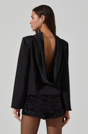 A woman is wearing a black satin blazer with a draped, open-back design. She has paired it with black sequin shorts and is accessorized with gold hoop earrings and a gold chain necklace with a dangling pendant.