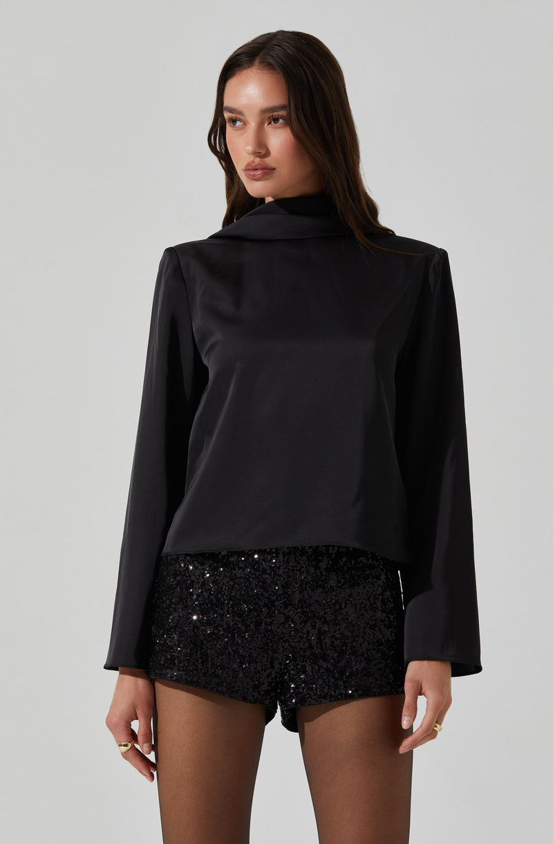 A woman wearing a black satin blazer with long sleeves and a notched lapel collar, paired with black sequin shorts, and sheer black tights. She has long, dark hair and is standing against a plain, light-colored background.