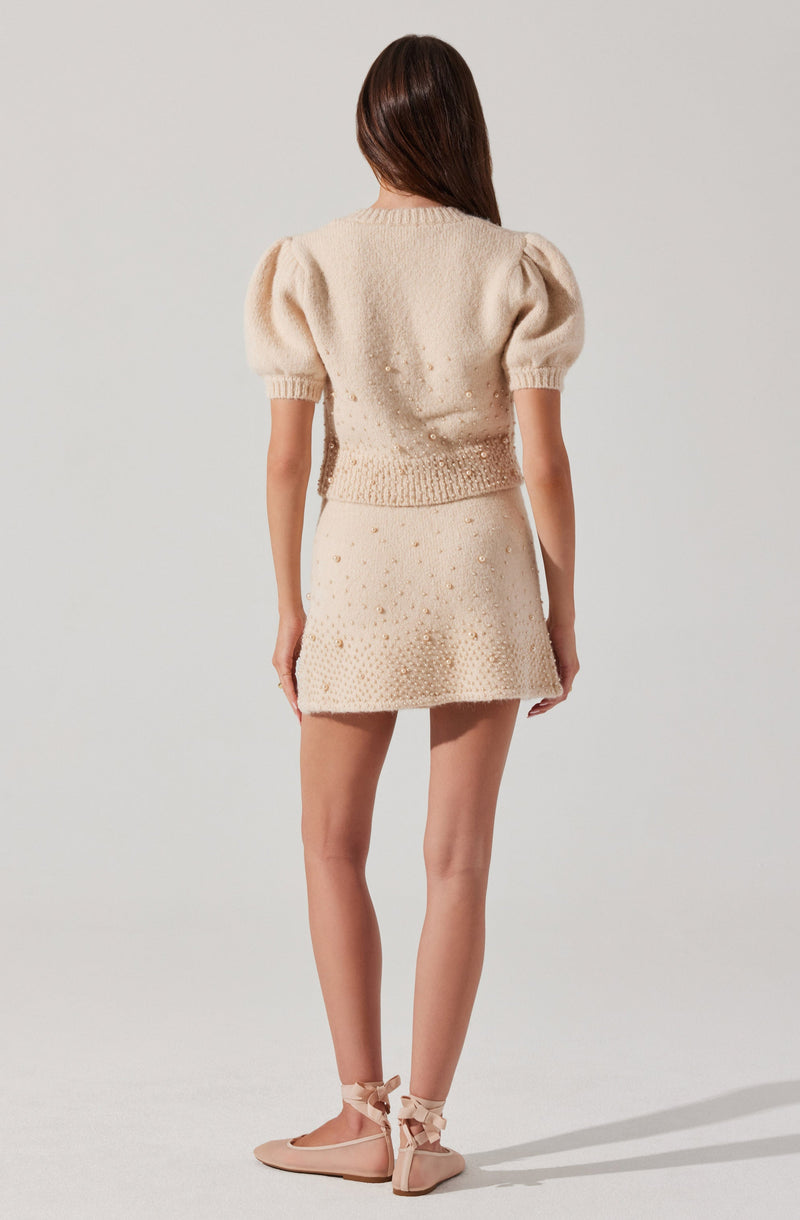 A woman is wearing a cream-colored, knitted two-piece outfit consisting of a short-sleeve top with puffed sleeves and a matching mini skirt. The top has a ribbed neckline and hem, while the skirt features a textured pattern with small decorative elements. She is also wearing beige flat shoes with ankle ties.
