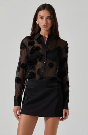 A young woman is wearing a black, sheer, long-sleeved button-up shirt with floral embellishments, paired with a black mini skirt. The shirt features embroidered or appliqué floral designs and has a classic collar with cuffs. The mini skirt is simple, with a tailored fit and subtle pocket details.