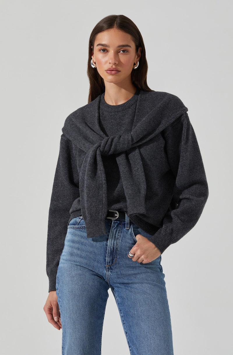 A woman wearing a dark gray sweater with a matching gray sweater tied around her shoulders. She is also wearing light blue high-waisted jeans with a black belt.