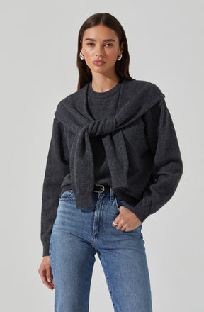 A woman wearing a dark gray sweater with a matching gray sweater tied around her shoulders. She is also wearing light blue high-waisted jeans with a black belt.