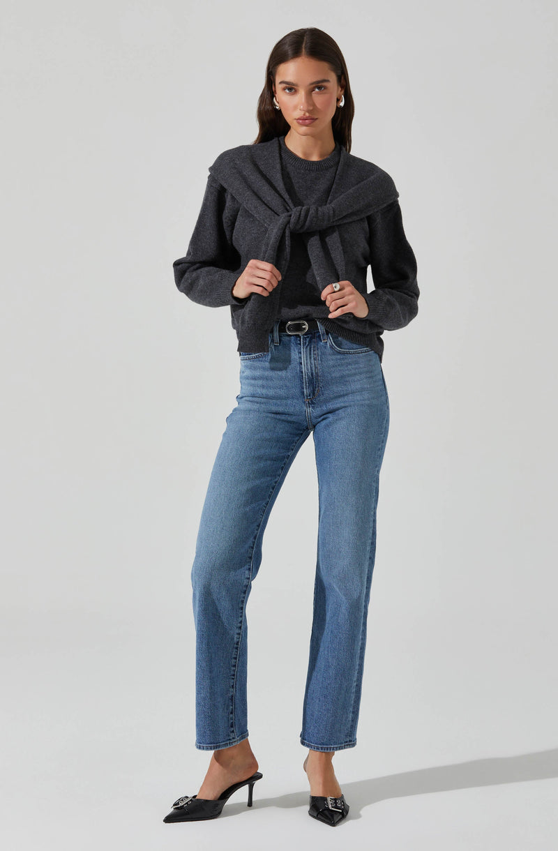 A woman is wearing a dark gray long-sleeved sweater tucked into light blue high-waisted jeans. She has a matching dark gray sweater tied around her shoulders. She is also wearing black pointed-toe high-heeled shoes with decorative embellishments on the front.