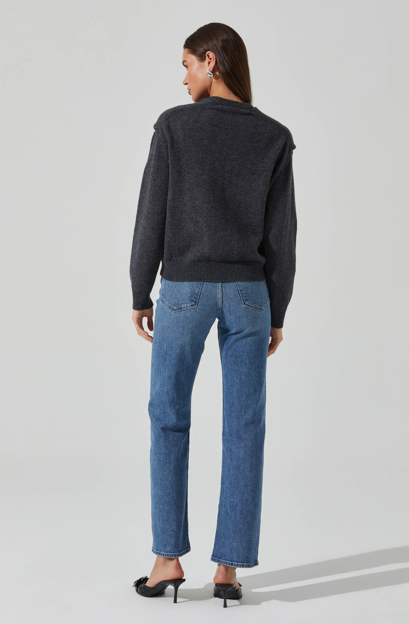 A woman is wearing a dark gray, long-sleeved knit sweater with a relaxed fit and ribbed cuffs and hem. She pairs it with light blue high-waisted jeans that have a slightly relaxed fit and classic back pockets. She completes her outfit with black, pointed-toe high-heeled shoes.