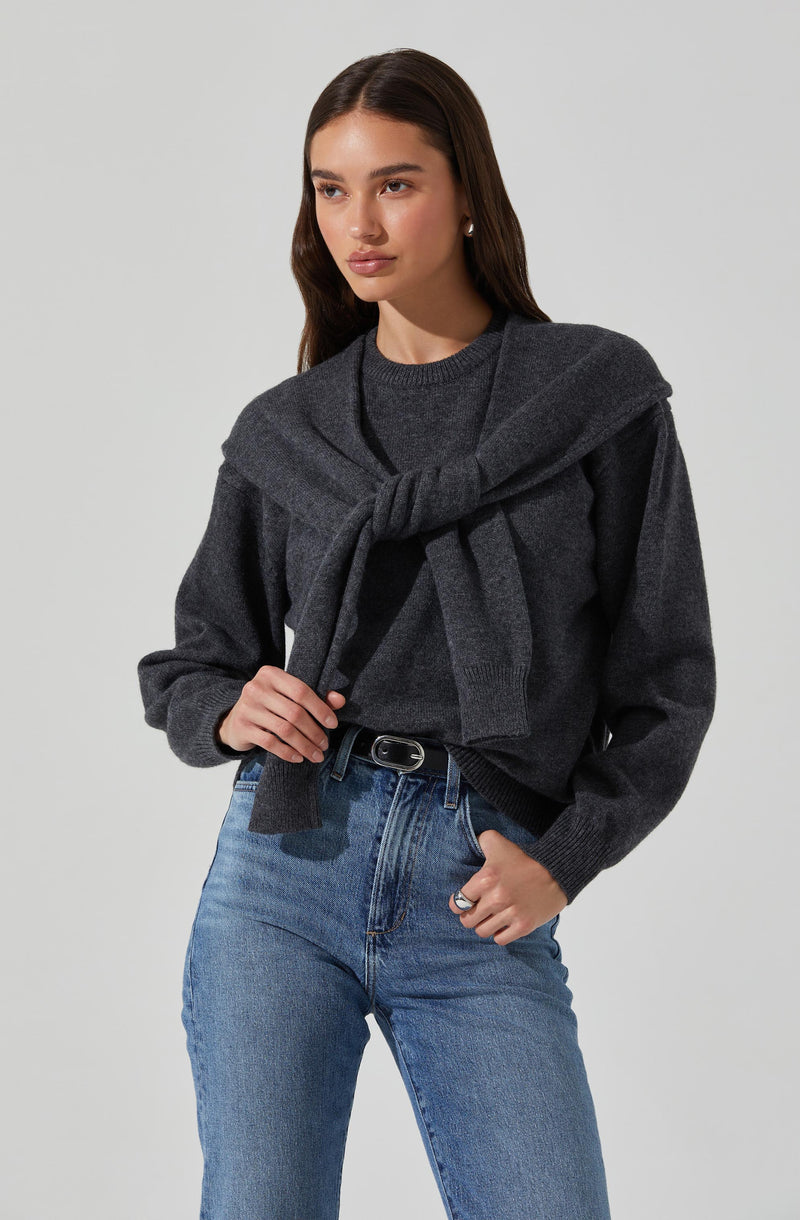 A young woman is dressed in a dark gray sweater with a crew neckline and long sleeves. She has a matching dark gray sweater draped over her shoulders, tied loosely in front. She is also wearing light blue high-waisted jeans with a black belt.