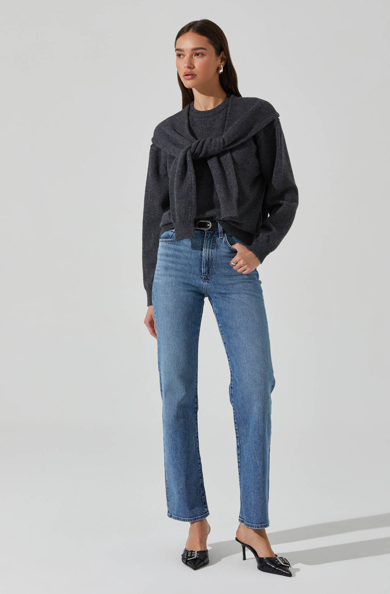 A woman is wearing a dark gray sweater with a matching gray sweater tied around her shoulders. She is also wearing light blue high-waisted jeans and black high-heeled shoes with a pointed toe and bow detail.