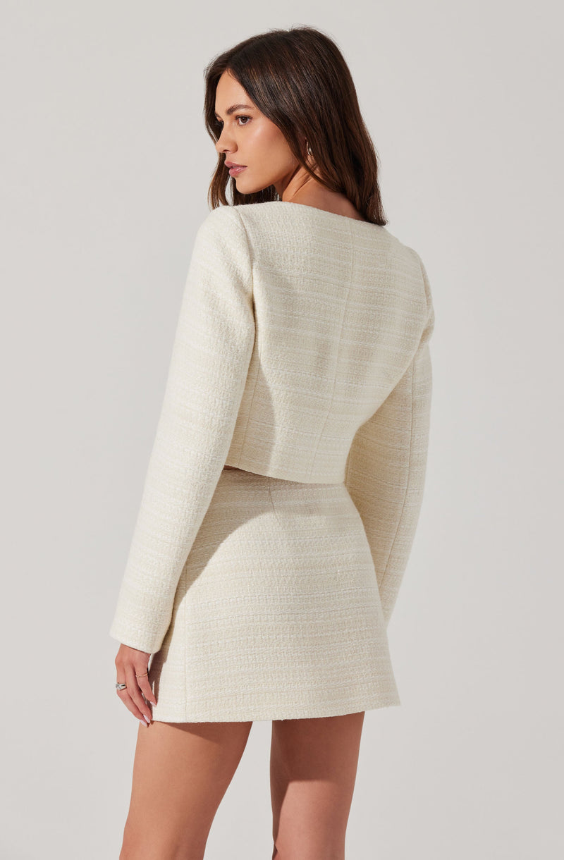 A woman is wearing a cream-colored, textured two-piece outfit consisting of a cropped blazer with long sleeves and a matching high-waisted, A-line mini skirt. The fabric of the outfit appears to have a subtle checkered or woven pattern.