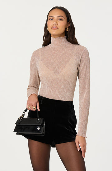 A woman is wearing a light pink, shimmery turtleneck sweater with long sleeves. She is also wearing black high-waisted shorts and sheer black tights. She has gold hoop earrings, multiple rings, and is holding a small black handbag with a top handle.