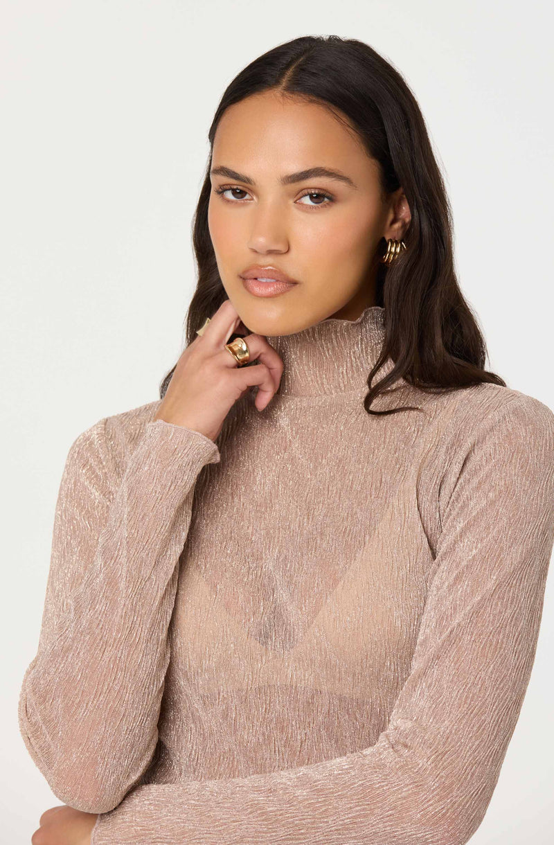 A woman with medium skin tone and dark wavy hair is wearing a high-neck, long-sleeved, shimmery beige top. She has gold earrings and a gold ring on her right hand.