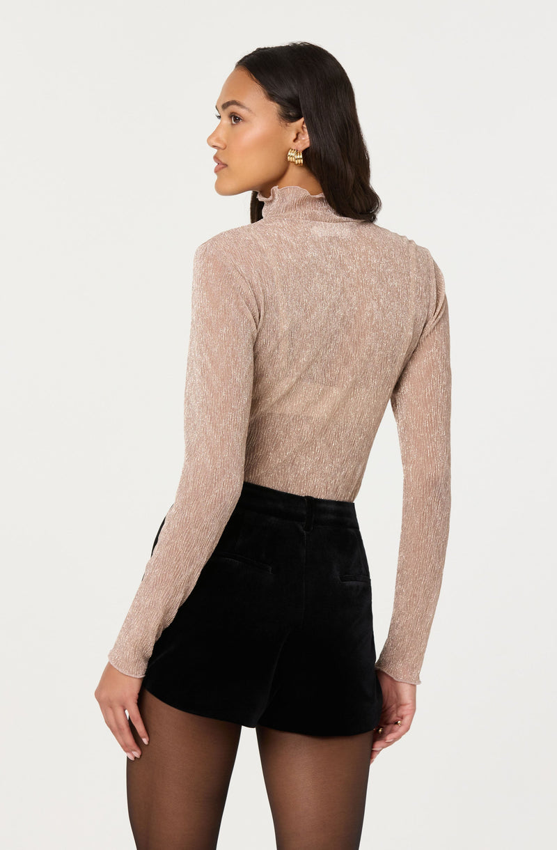 A woman is wearing a fitted, long-sleeve, beige turtleneck sweater with a subtle shimmer and a ruffled neckline. She pairs it with high-waisted black shorts and sheer black tights. The woman has dark, wavy hair and is accessorized with large, gold textured earrings.
