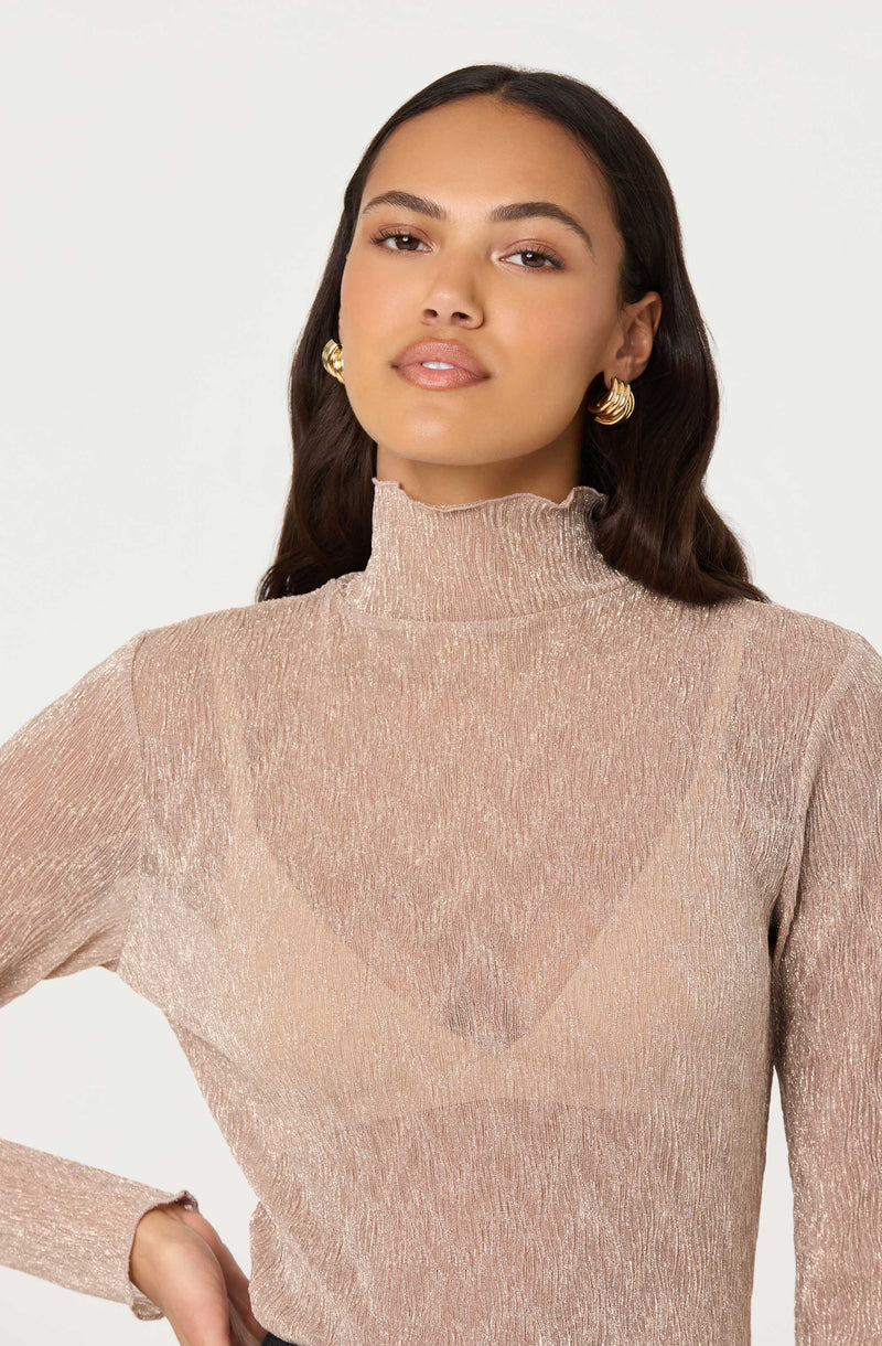 A woman is wearing a shimmery, long-sleeved, high-neck blouse in a soft, metallic blush color. The fabric has a textured, slightly crinkled appearance with a subtle sheen.