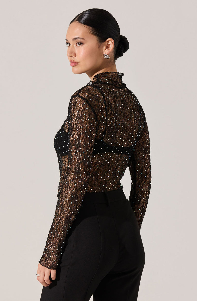 A woman is wearing a sheer black long-sleeve top with a high ruffled collar. The top is decorated with small, shiny, white polka dots. She pairs it with high-waisted black pants.