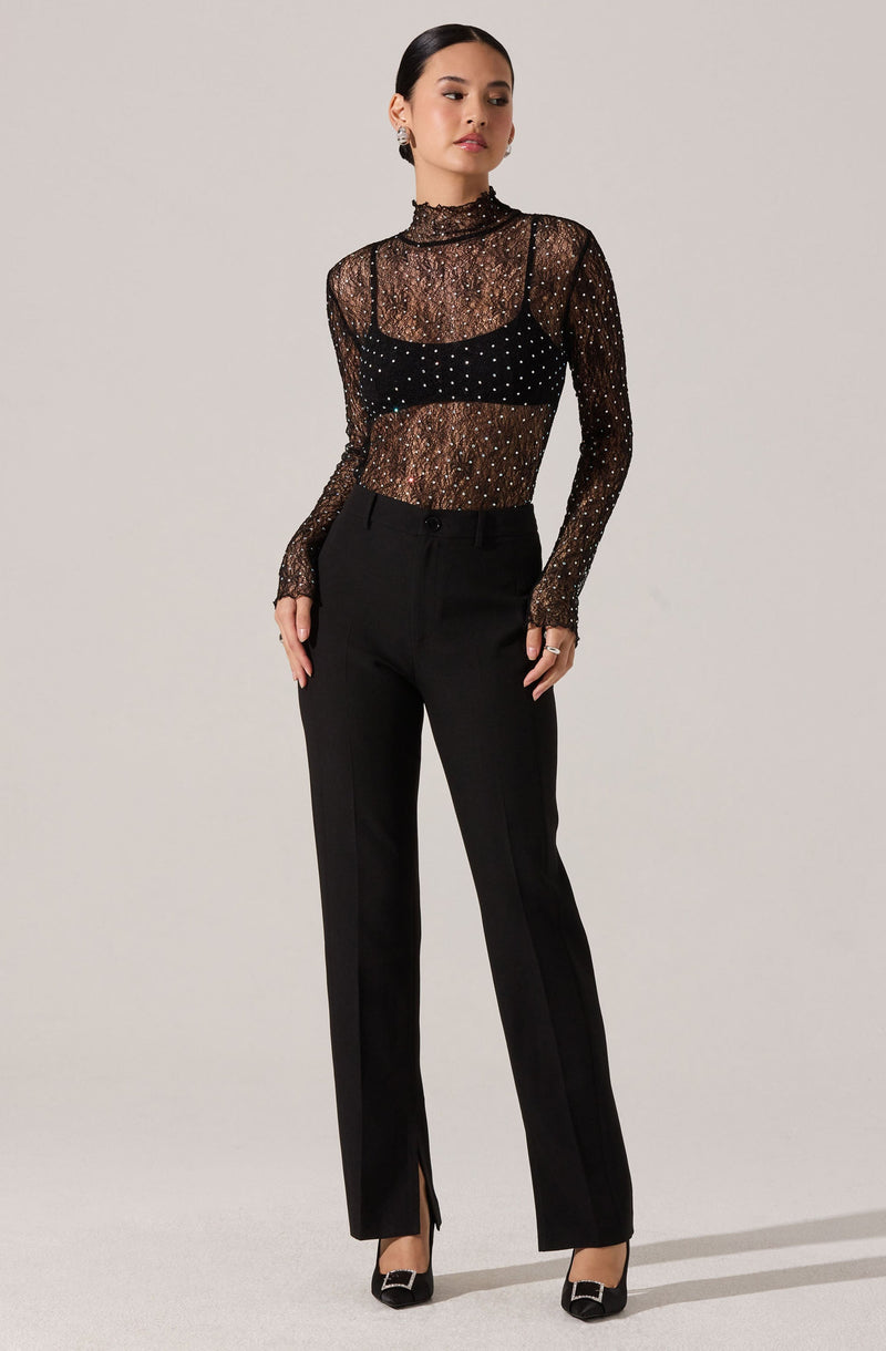 A woman is wearing a sheer black long-sleeve top with a high neckline, decorated with small, shiny embellishments. Underneath, she has a black bralette that is visible through the sheer fabric. She is also wearing high-waisted black trousers with a straight-leg fit, featuring a zipper and button closure, and small zippers at the hem. She completes the look with black heels that have a decorative buckle on the front.