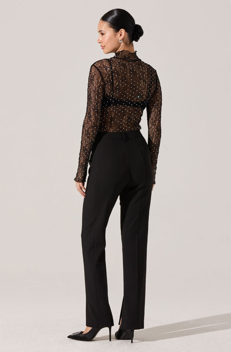 A woman is wearing a sheer, long-sleeved black blouse adorned with numerous small, shiny embellishments. She pairs this with high-waisted black trousers that have a tailored fit and a slight flare at the bottom. She is also wearing black high-heeled shoes with pointed toes.