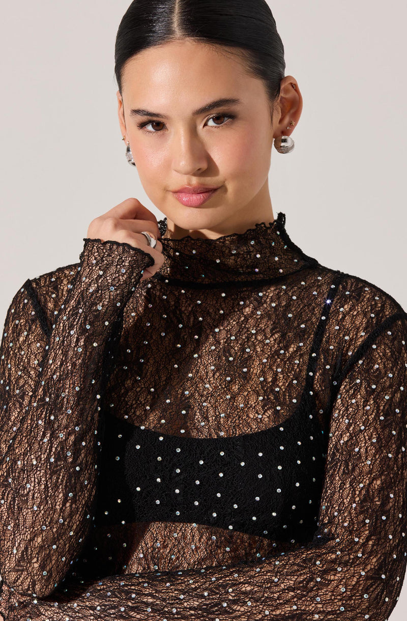 The woman is wearing a sheer black lace top with long sleeves and a high neckline, adorned with small, evenly spaced rhinestones. Underneath, she has a black camisole with thin straps and a pattern of rhinestones that complement the lace top. She is accessorized with silver earrings and a ring.