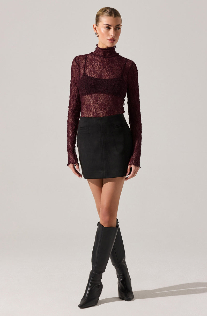 A woman is wearing a long-sleeved, burgundy lace turtleneck top that is semi-sheer, paired with a high-waisted black mini skirt. She is also wearing knee-high black boots with a slight heel.
