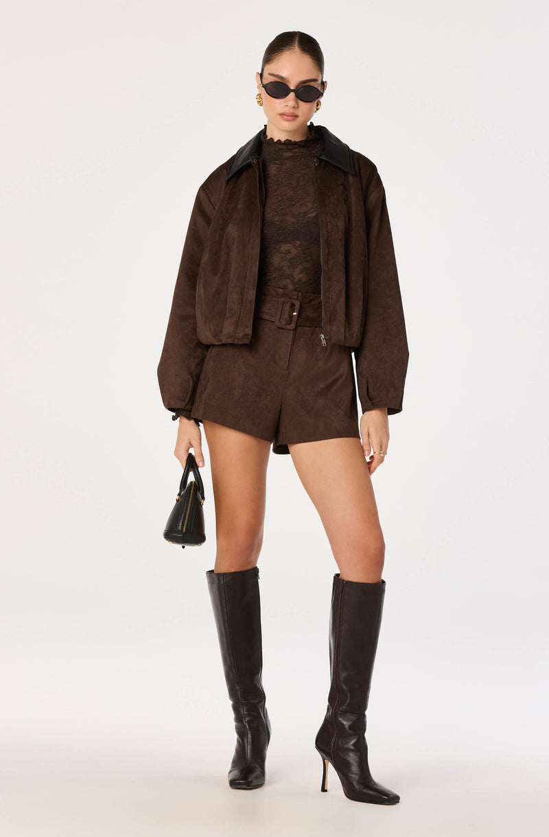Woman in brown suede jacket, matching shorts, black lace top, knee-high black boots, and sunglasses.
