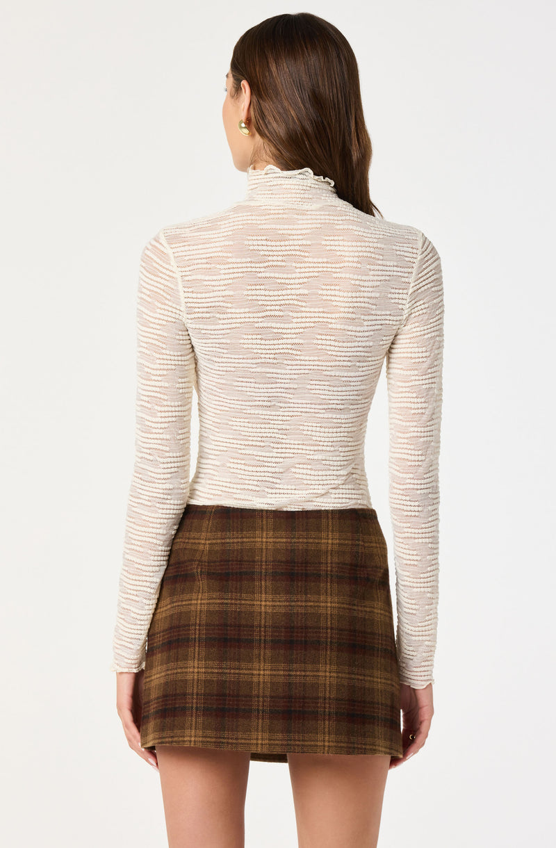 Cream ribbed turtleneck sweater with ruffled collar and long sleeves, paired with a brown plaid skirt.