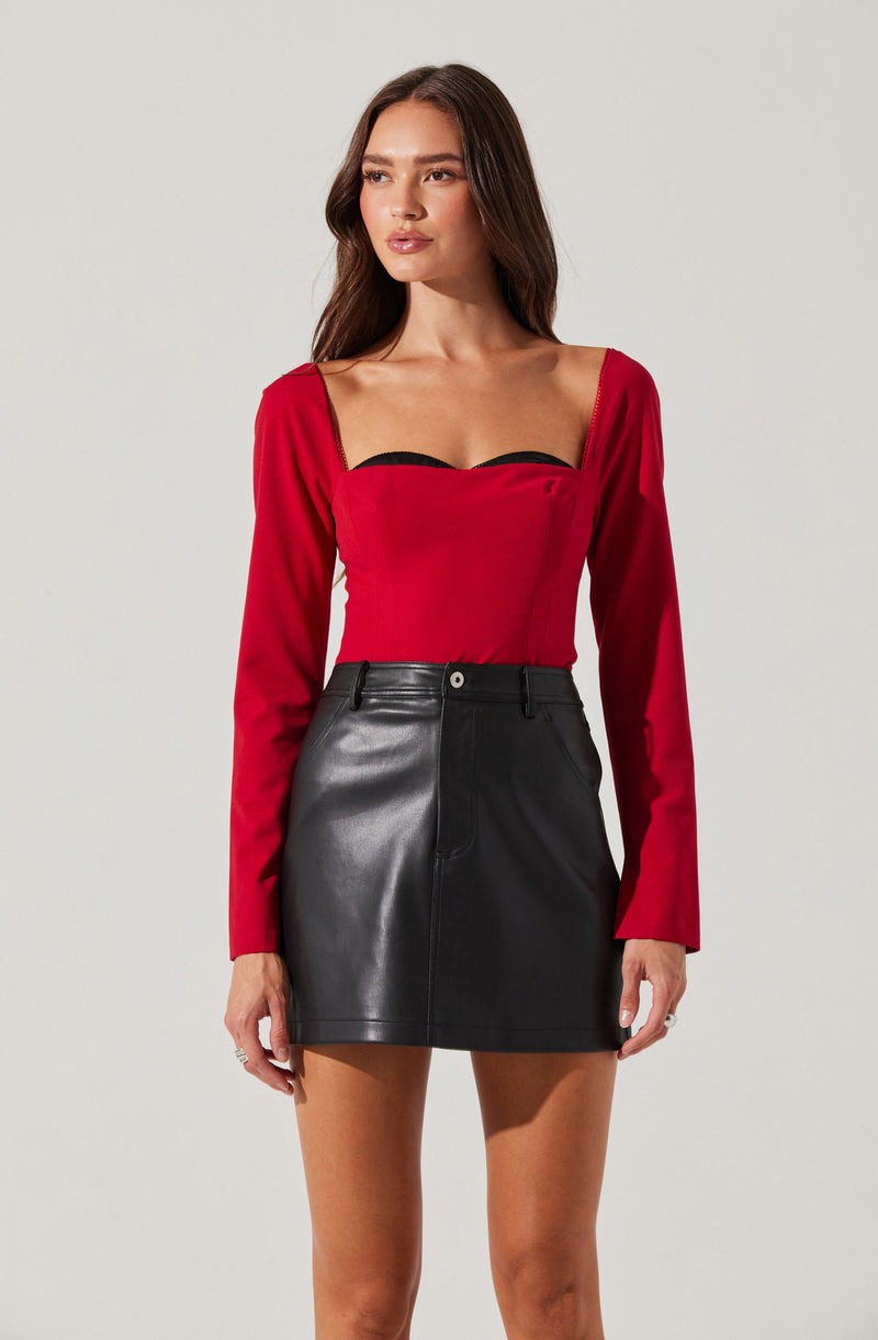 A woman is wearing a fitted red long-sleeve top with a wide, square neckline and black trim details along the bust. She pairs it with a high-waisted black leather mini skirt with front seams and belt loops.