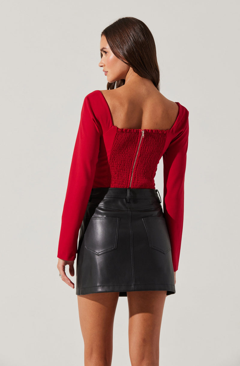 A woman is wearing a red long-sleeve top with ruching and a front zipper, paired with a black leather mini skirt featuring back pockets and visible stitching.
