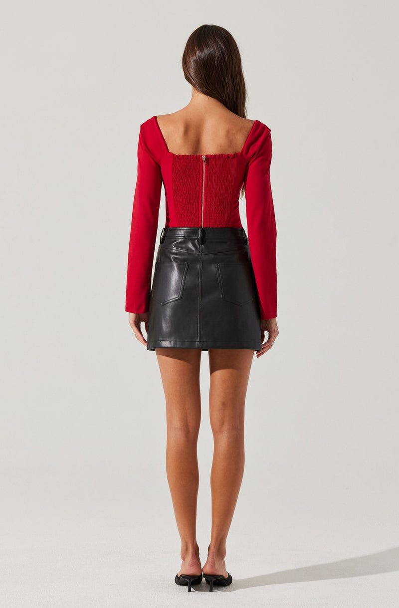 A woman is wearing a red long-sleeve top with a gathered back and a central zipper, paired with a high-waisted black leather mini skirt with two back pockets. She is also wearing black open-toe heels.