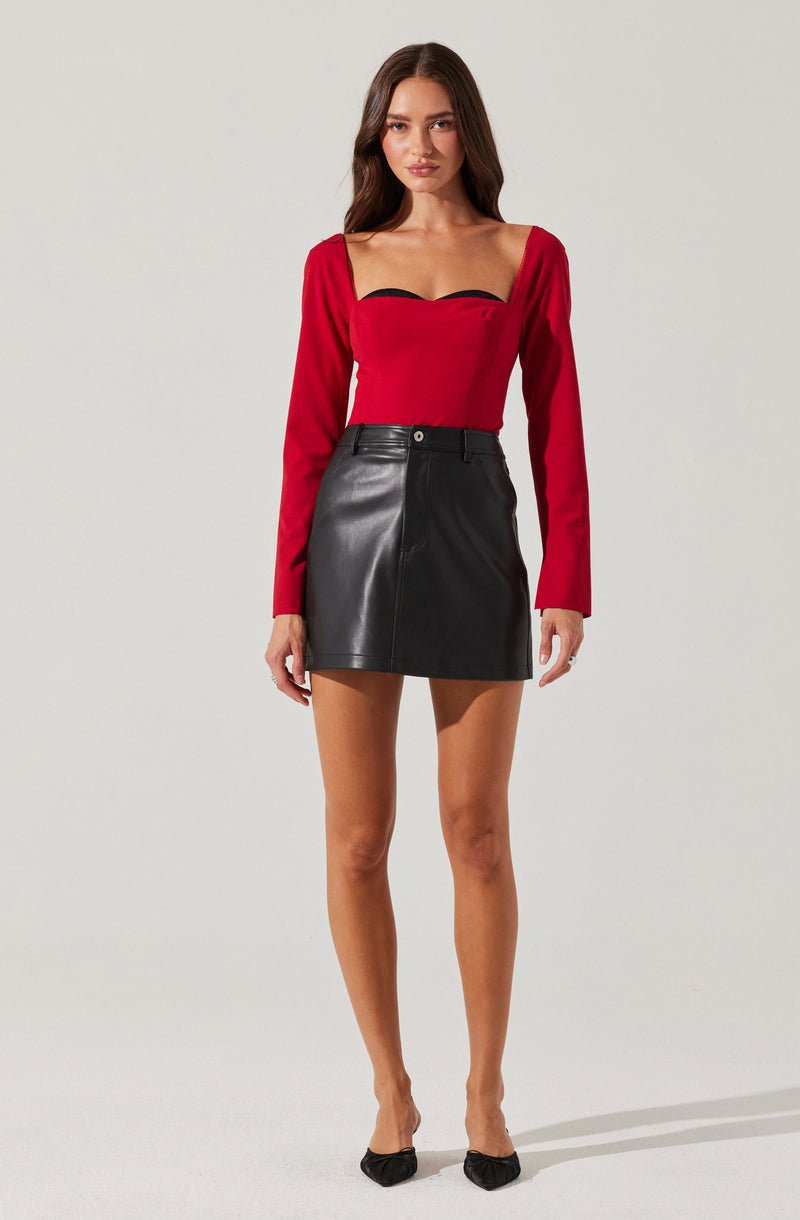 A woman is wearing a red long-sleeve top with a sweetheart neckline accented by black trim, paired with a high-waisted black leather mini skirt. She is also wearing black pointed-toe shoes with embellishments.