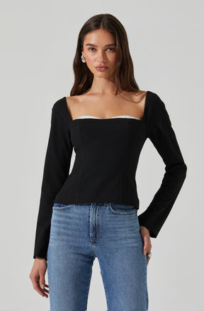 A woman is wearing a black long-sleeve top with a fitted bustier style bodice, featuring a sweetheart neckline and subtle shoulder seam details. The top is paired with light blue high-waisted jeans.