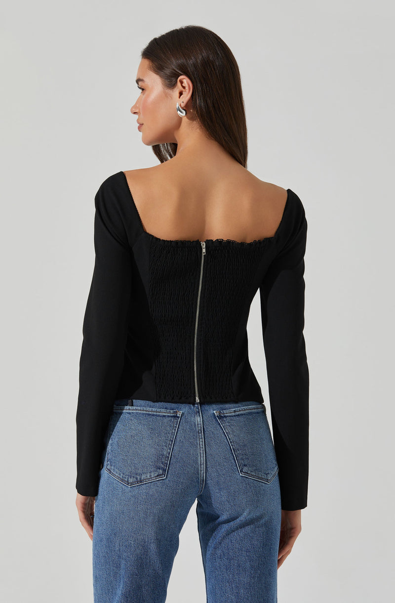 A woman is wearing a black long-sleeve top with a wide, square neckline and a central zipper detail running down the back. The top has a fitted, ruched texture on the back panel. She is also wearing high-waisted blue jeans with visible stitching on the pockets and seams.